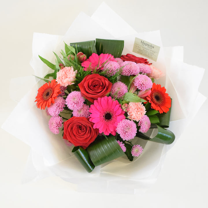 the Buckingham Bouquet from Magnolia the Florist,  features a stunning combination of red and pink-toned flowers, including roses, gerbera, alstroemeria, carnation, chrysanthemum, and foliage