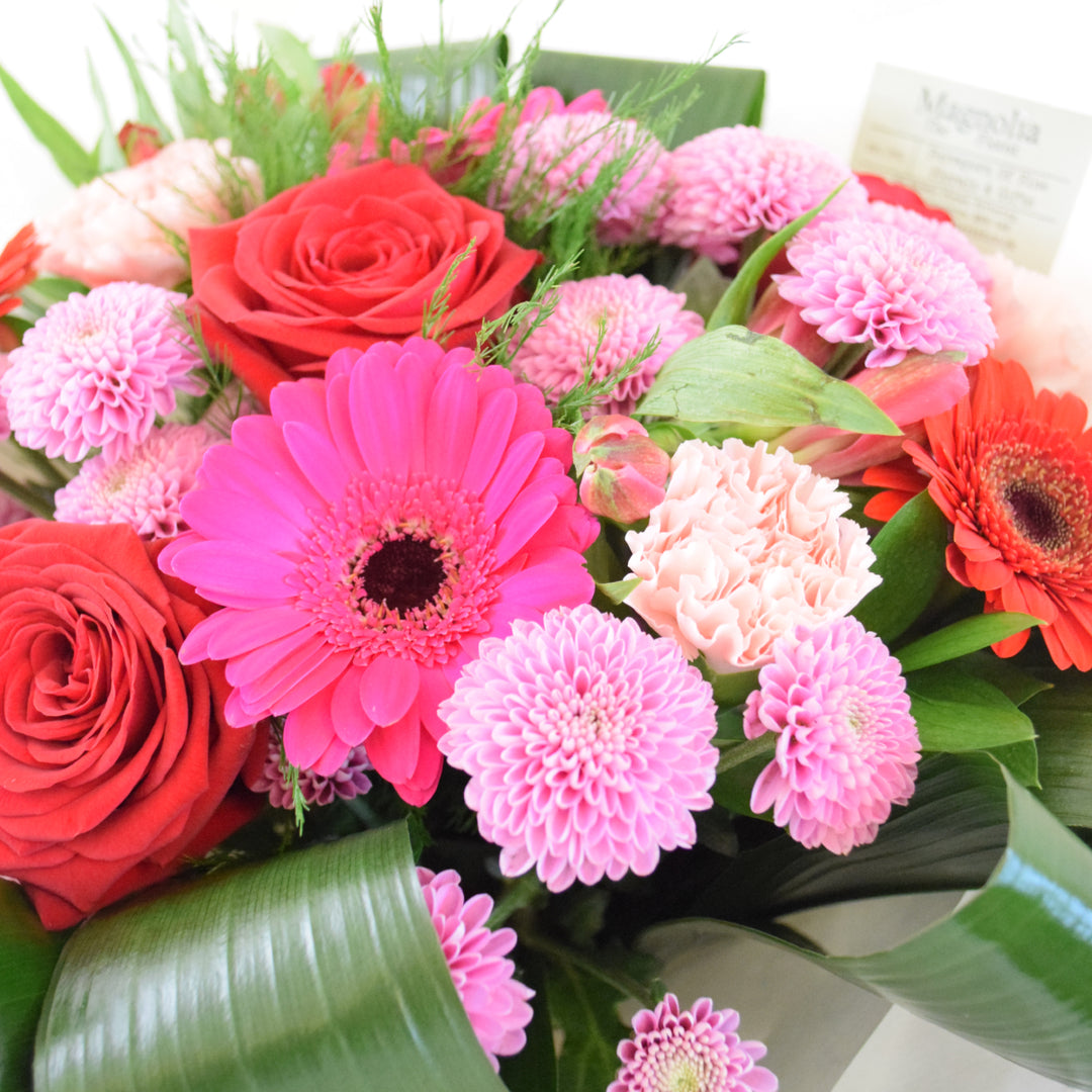the Buckingham Bouquet from Magnolia the Florist,  features a stunning combination of red and pink-toned flowers, including roses, gerbera, alstroemeria, carnation, chrysanthemum, and foliage