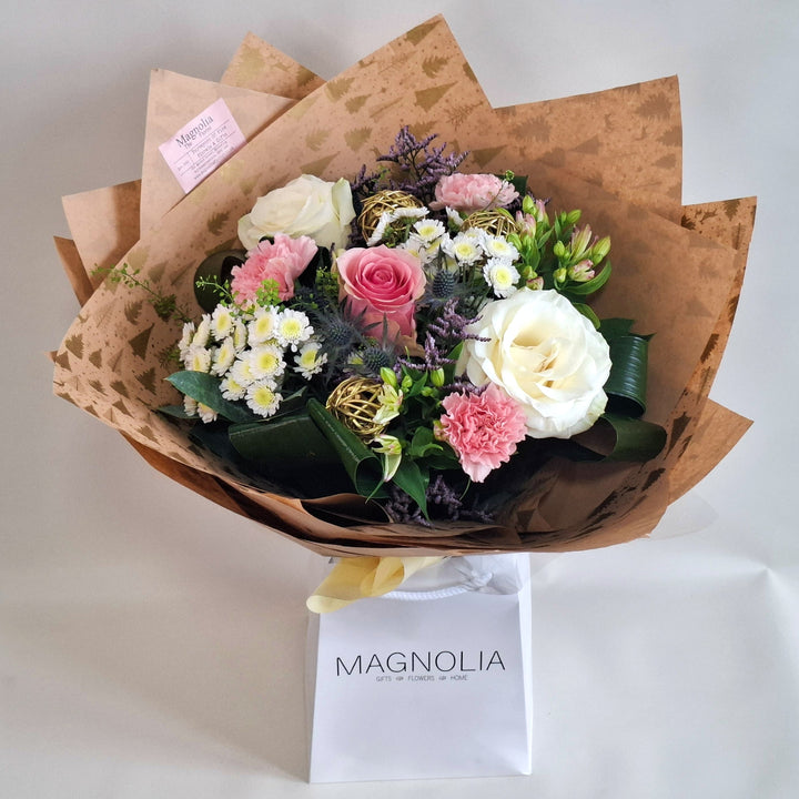 Bouquet of pink and white toned flowers wrapped in kraft paper with a 'MAGNOLIA' branded white bag underneath.