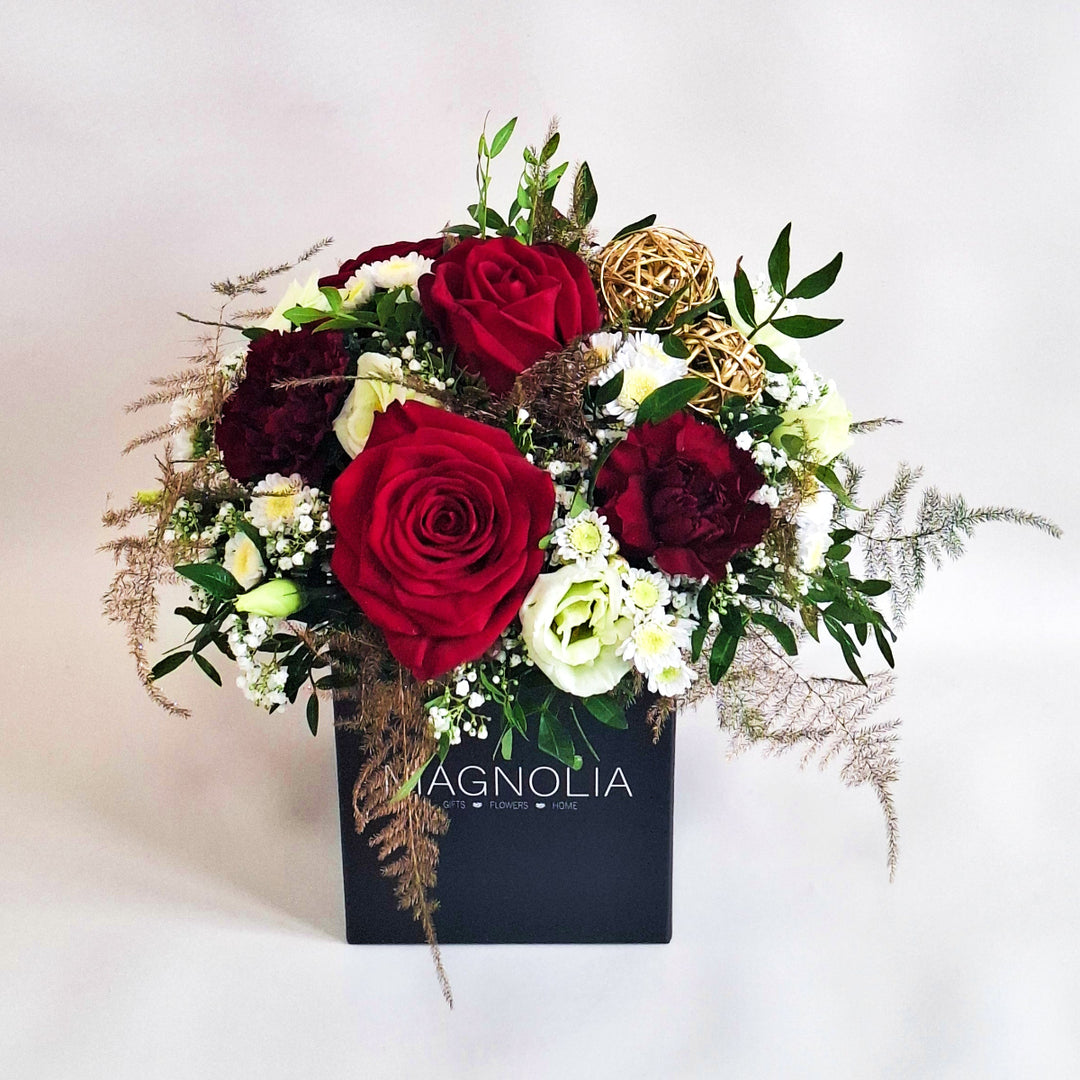 Floral arrangement with red and burgundy roses, greenery, and decorative elements in a black box.