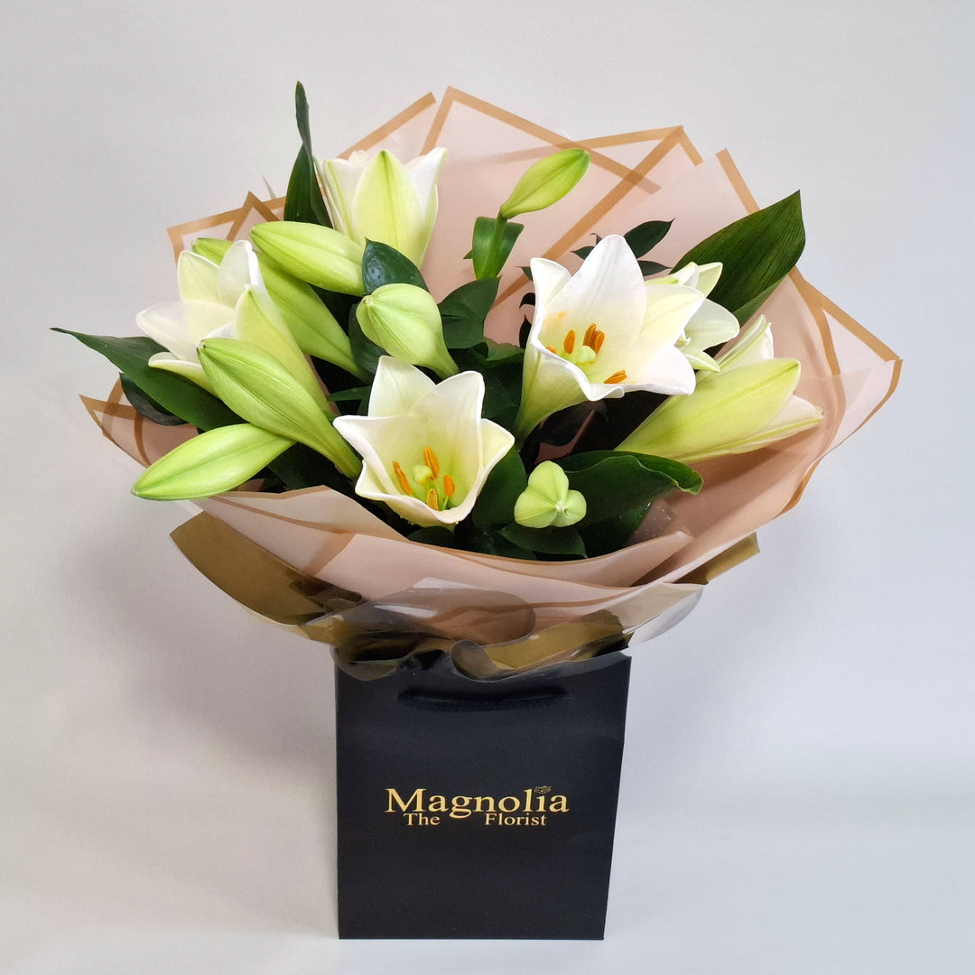 Balmoral Bouquet - Magnolia the Florist A tall, sculptural hand-tied bouquet built around elegant lilies, aspidistra and ruscus. The bouquet is finished in an aqua bubble and then presented in one of our Magnolia-branded hand-tied carriers. For scale, our petite size measures approximately 50cm tall and 38cm wide. Larger sizes increase in overall width while maintaining the same balanced proportions. Please note this item requires 1-day notice to order.  1