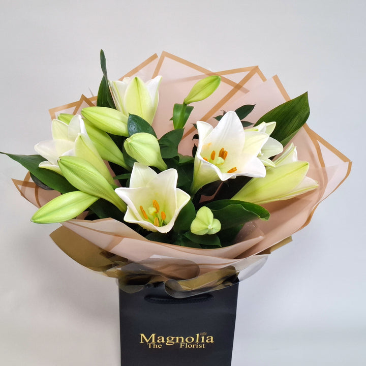 Balmoral Bouquet - Magnolia the Florist A tall, sculptural hand-tied bouquet built around elegant lilies, aspidistra and ruscus. The bouquet is finished in an aqua bubble and then presented in one of our Magnolia-branded hand-tied carriers. For scale, our petite size measures approximately 50cm tall and 38cm wide. Larger sizes increase in overall width while maintaining the same balanced proportions. Please note this item requires 1-day notice to order.  3