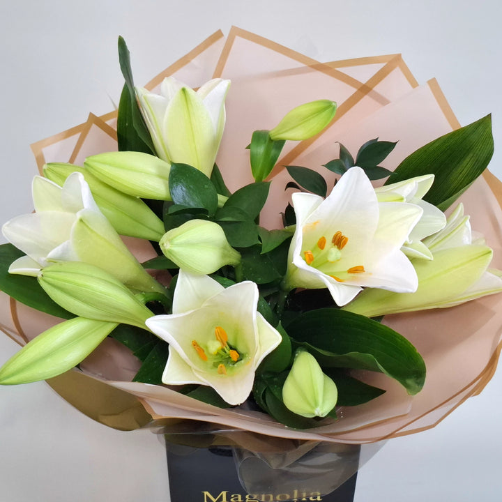 Balmoral Bouquet - Magnolia the Florist A tall, sculptural hand-tied bouquet built around elegant lilies, aspidistra and ruscus. The bouquet is finished in an aqua bubble and then presented in one of our Magnolia-branded hand-tied carriers. For scale, our petite size measures approximately 50cm tall and 38cm wide. Larger sizes increase in overall width while maintaining the same balanced proportions. Please note this item requires 1-day notice to order.  2