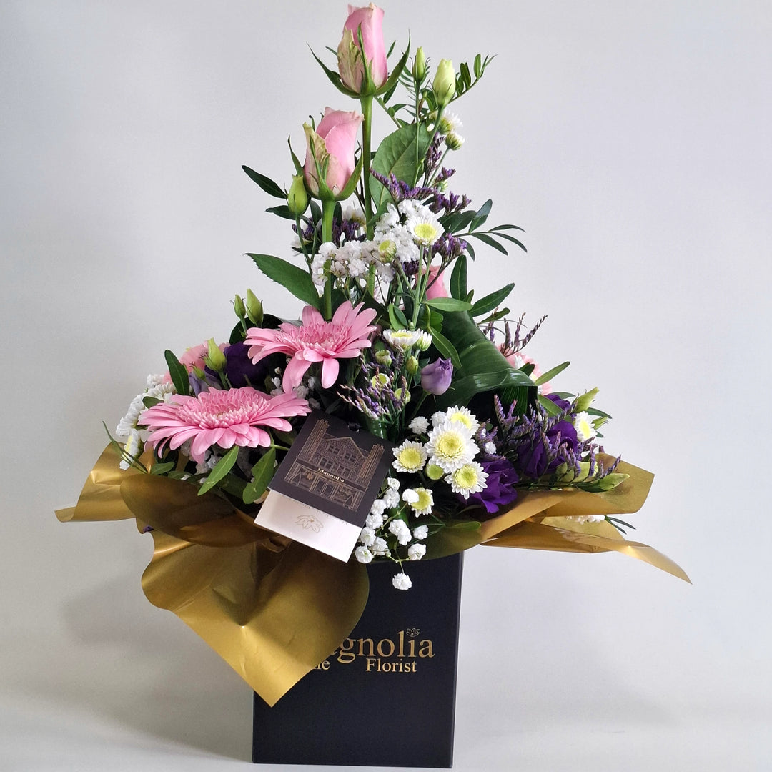 Islay Arrangement - Magnolia the Florist Islay is a soft, feminine floral arrangement crafted with pink roses, pink carnations, pink germini, white chrysanthemum, purple lisianthus, folded aspidistra, limonium, gyp and pistache. Presented in our Magnolia‑branded box and arranged in floral foam for long‑lasting freshness. For scale purposes, our branded box is 15cm (h) x 12cm (w) x 12cm (d), giving an overall size of approximately 35cm (h) x 18cm (w).  1