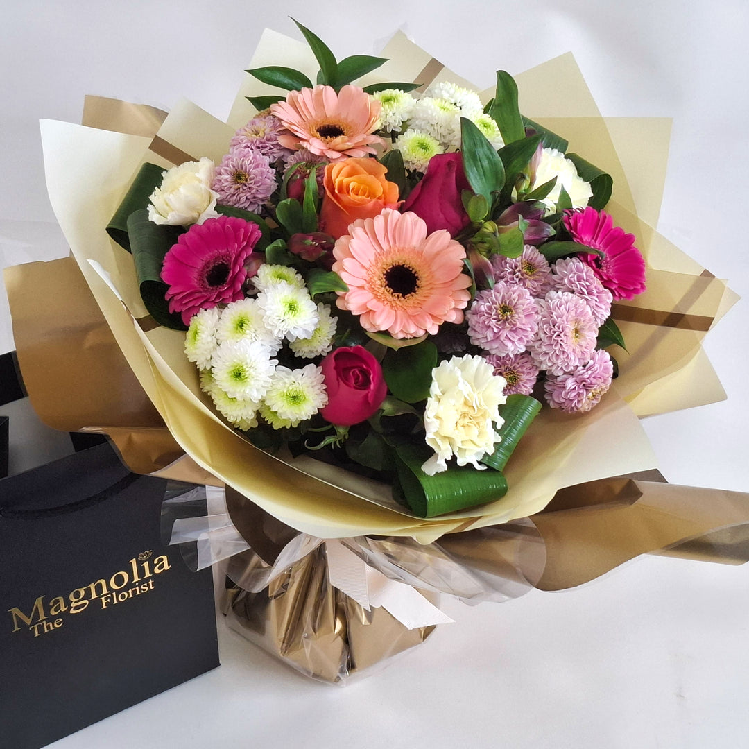 Joy For Mum Hand-tied Bouquet - Magnolia the Florist Celebrate Mother's Day with this stunning hand-tied bouquet crafted especially for mum. A blend of roses, germini, carnations, and chrysanthemums in cerise, peach, pink, and white tones creates a joyful arrangement that radiates warmth and appreciation. Presented in our signature Magnolia branded box bag, this bouquet arrives fresh and ready to brighten her day with elegance and charm.  3