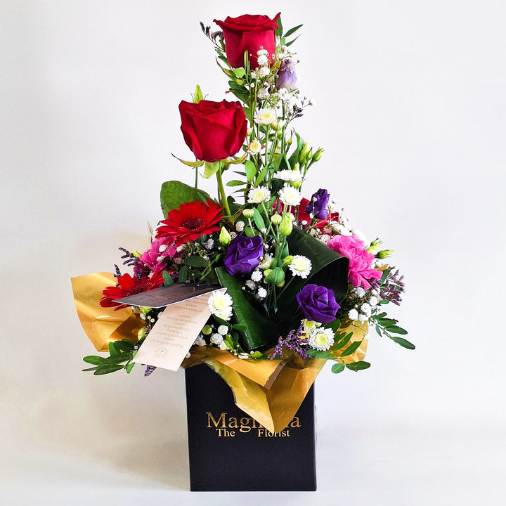 Jura Arrangement - Magnolia the Florist Jura is a dramatic, structured floral arrangement featuring red germini, red roses, cerise carnations, white chrysanthemum, purple lisianthus, folded aspidistra, limonium, gyp and pistache. Presented in our Magnolia‑branded box and arranged in floral foam for long‑lasting freshness. For scale purposes, our branded box is 15cm (h) x 12cm (w) x 12cm (d), giving an overall size of approximately 35cm (h) x 18cm (w).  1