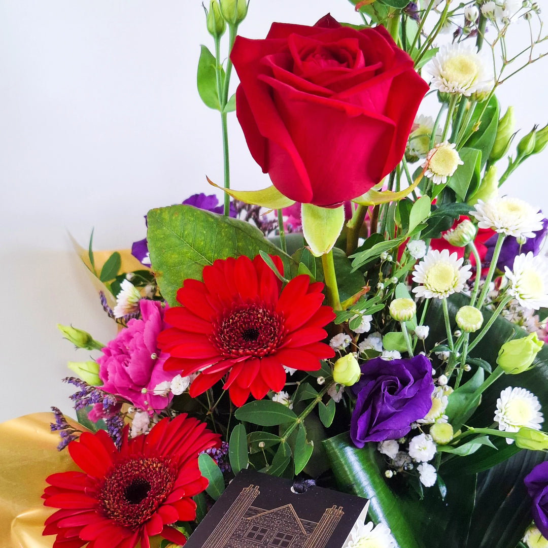 Jura Arrangement - Magnolia the Florist Jura is a dramatic, structured floral arrangement featuring red germini, red roses, cerise carnations, white chrysanthemum, purple lisianthus, folded aspidistra, limonium, gyp and pistache. Presented in our Magnolia‑branded box and arranged in floral foam for long‑lasting freshness. For scale purposes, our branded box is 15cm (h) x 12cm (w) x 12cm (d), giving an overall size of approximately 35cm (h) x 18cm (w).  2
