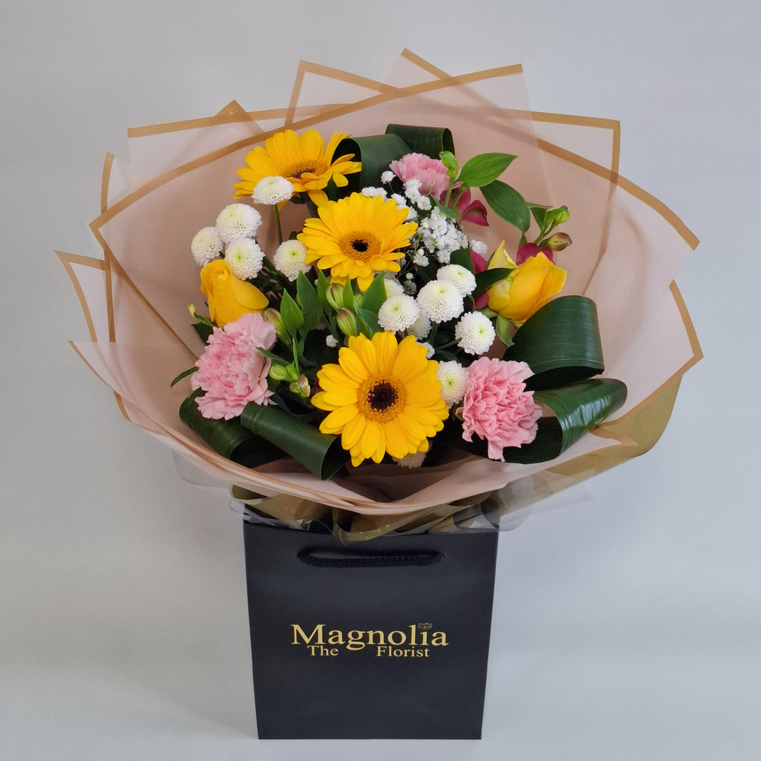 Lilford Bouquet - Magnolia the Florist A warm, sunny hand‑tied bouquet featuring yellow germini, pink bloom carnations, yellow bloom roses, pink alstroemeria, white spray chrysanthemums and gyp. The bouquet is finished in an aqua bubble and then presented in one of our Magnolia‑branded hand‑tied carriers. For scale, our petite size measures approximately 50cm tall and 38cm wide. Larger sizes increase in overall width while maintaining the same balanced proportions.  1