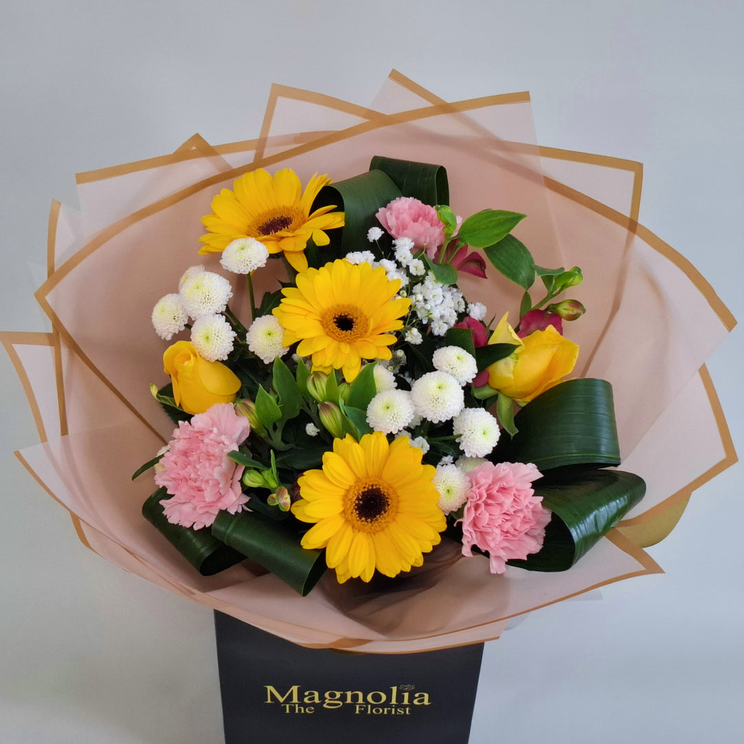 Lilford Bouquet - Magnolia the Florist A warm, sunny hand‑tied bouquet featuring yellow germini, pink bloom carnations, yellow bloom roses, pink alstroemeria, white spray chrysanthemums and gyp. The bouquet is finished in an aqua bubble and then presented in one of our Magnolia‑branded hand‑tied carriers. For scale, our petite size measures approximately 50cm tall and 38cm wide. Larger sizes increase in overall width while maintaining the same balanced proportions.  3