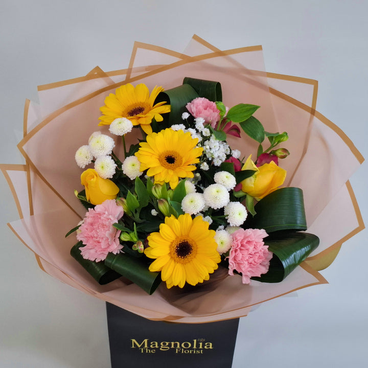 Lilford Bouquet - Magnolia the Florist A warm, sunny hand‑tied bouquet featuring yellow germini, pink bloom carnations, yellow bloom roses, pink alstroemeria, white spray chrysanthemums and gyp. The bouquet is finished in an aqua bubble and then presented in one of our Magnolia‑branded hand‑tied carriers. For scale, our petite size measures approximately 50cm tall and 38cm wide. Larger sizes increase in overall width while maintaining the same balanced proportions.  3