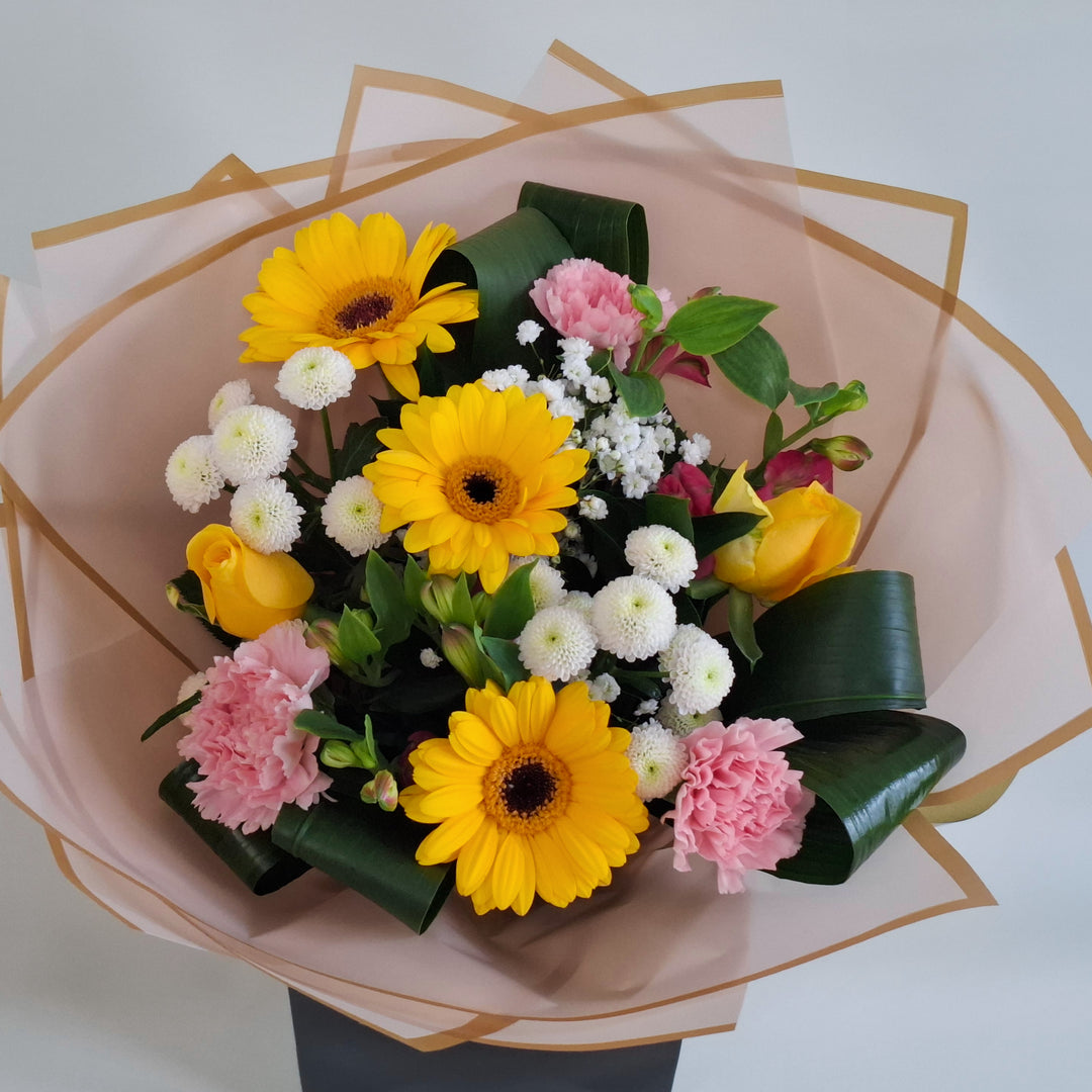 Lilford Bouquet - Magnolia the Florist A warm, sunny hand‑tied bouquet featuring yellow germini, pink bloom carnations, yellow bloom roses, pink alstroemeria, white spray chrysanthemums and gyp. The bouquet is finished in an aqua bubble and then presented in one of our Magnolia‑branded hand‑tied carriers. For scale, our petite size measures approximately 50cm tall and 38cm wide. Larger sizes increase in overall width while maintaining the same balanced proportions.  2