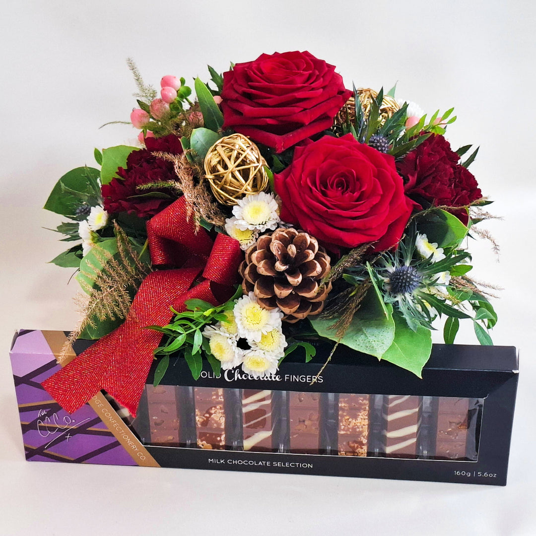 Floral arrangement with red roses and decorative elements, featuring a box of chocolate fingers.