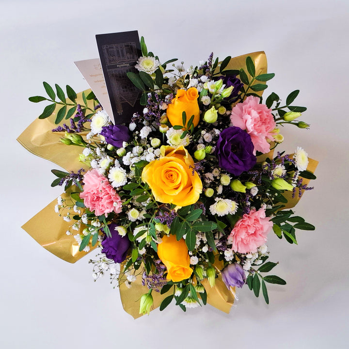 Dainty Somerset Arrangement - Magnolia the Florist A bright and uplifting floral arrangement designed to bring warmth into any space. Somerset features a joyful blend of yellow roses, pink carnations, white chrysanthemum, purple lisianthus, limonium, gyp and pistache. Presented in our Magnolia‑branded box and arranged in floral foam for long‑lasting freshness. For scale purposes, our branded box is 15cm(h) x 12cm(w) x 12cm(d), giving an overall size of approximately 25cm (h) x 18cm (w).  2