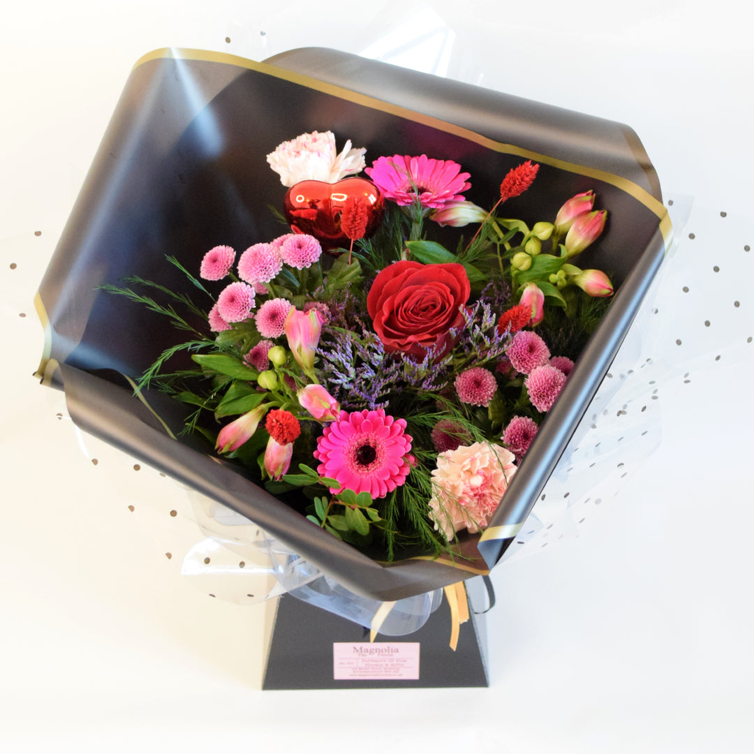 Sweet Heart Hand-tied Bouquet - Magnolia the Florist Indulge in the ultimate expression of love with our Sweet Heart Box. This luxurious hand-tied bouquet features a single red rose, cerise germini, pink chrysanthemums, pink alstroemeria, purple statice, and greenery, all expertly arranged and presented in one of our sleek black boxes. Treat yourself or a loved one to this elegant and exclusive gift. For scale purposes, this hand-tied is approx 42cm(h) x 25cm(w) for the Petite size.  1