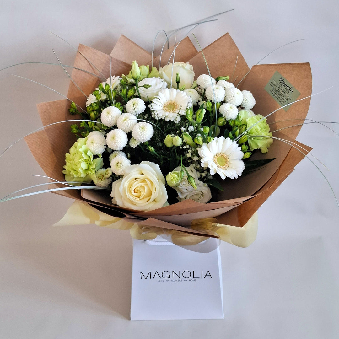 Bouquet of flowers wrapped in brown paper, in shades of white and Green and is presented a Magnolia the Florist Branded Bag.