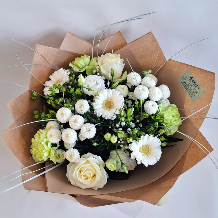 Bouquet of flowers wrapped in brown paper, in shades of white and Green and is presented a Magnolia the Florist Branded Bag.