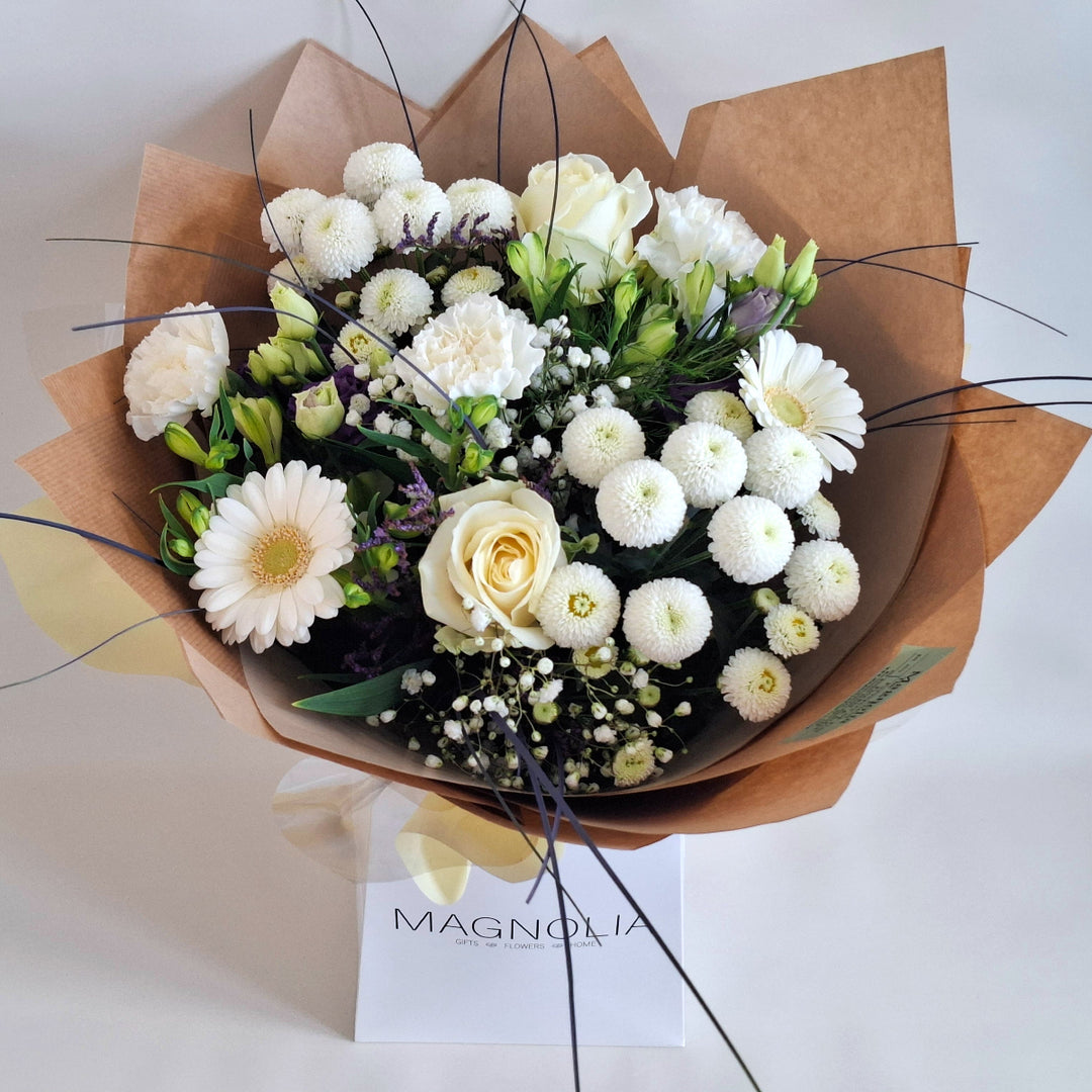 Bouquet of white and purple flowers wrapped in brown paper, presented in a Magnolia the Florist branded bag.