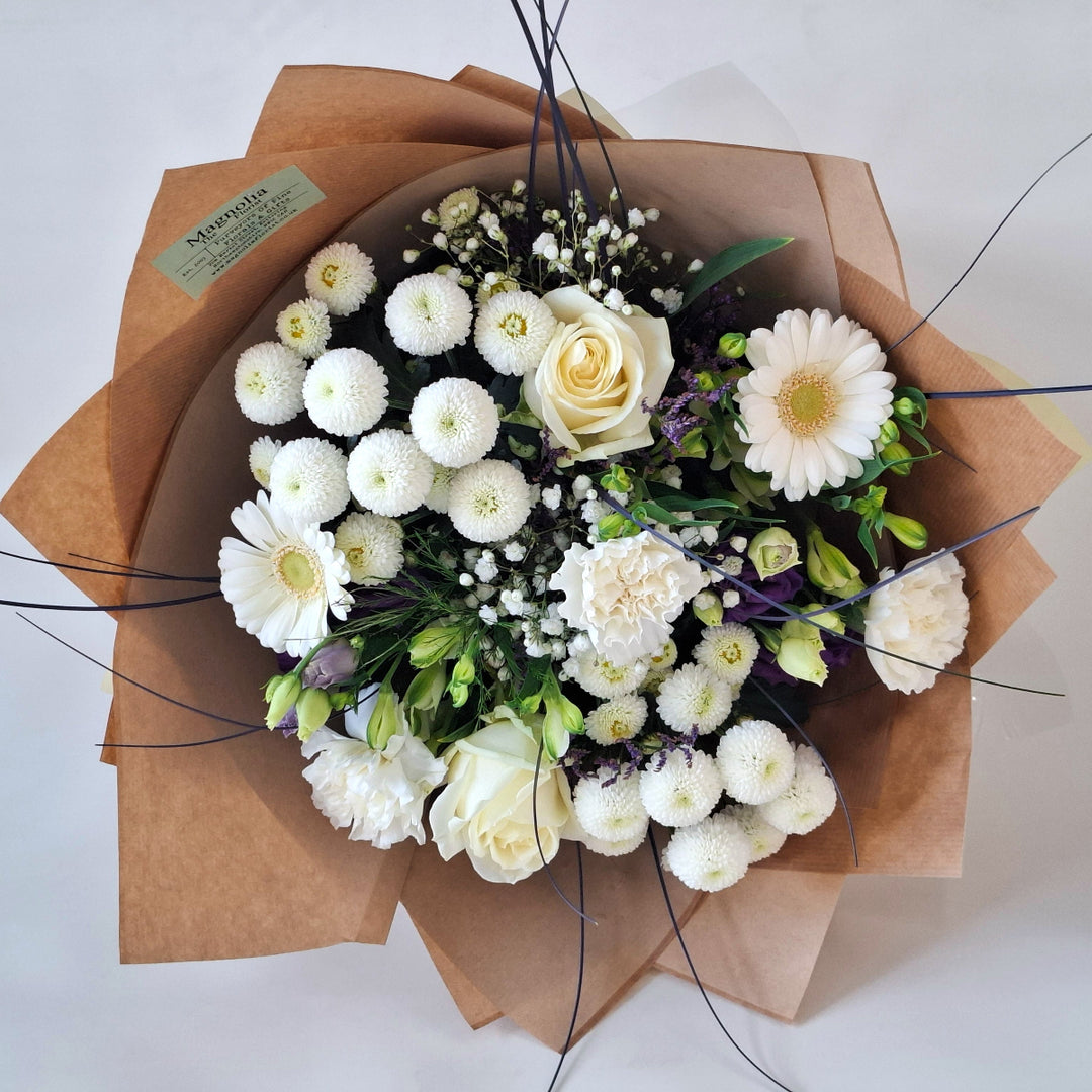 Bouquet of white and purple flowers wrapped in brown paper, presented in a Magnolia the Florist branded bag.