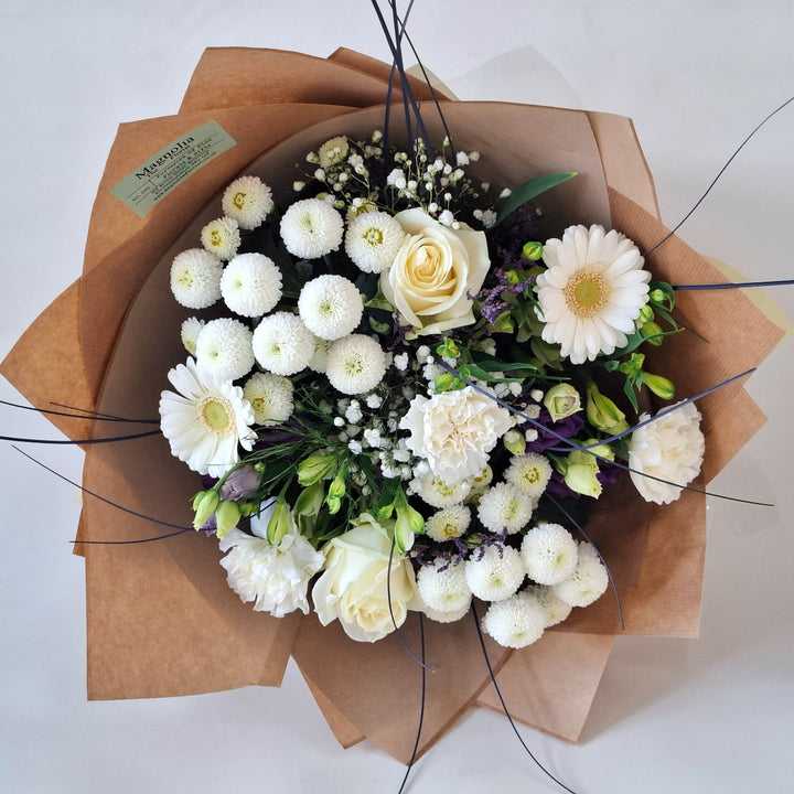 Bouquet of white and purple flowers wrapped in brown paper, presented in a Magnolia the Florist branded bag.