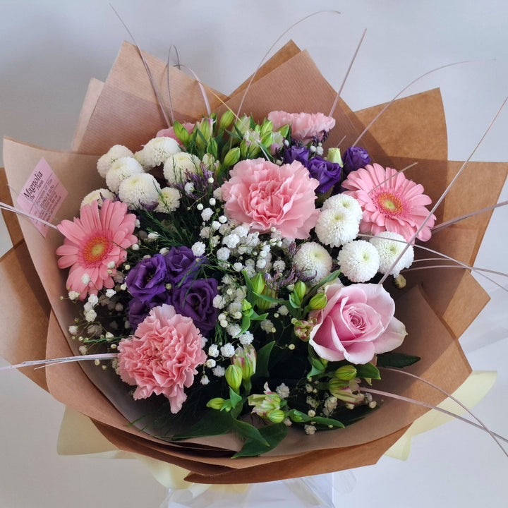 Bouquet of flowers wrapped in brown paper, in shade sof pink, white and purple and in a Magnolia the Florist Branded Bag.