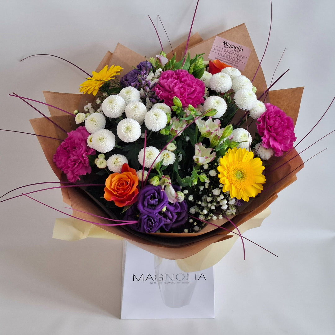 Bouquet of Vibrant flowers wrapped in brown paper, presented in a Magnolia the Florist branded bag.