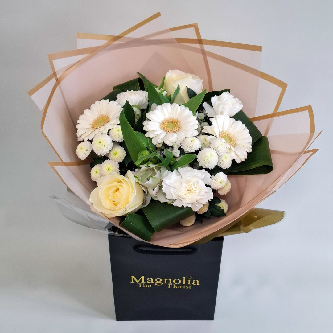 Wadenhoe Bouquet - Magnolia the Florist A pure white hand‑tied bouquet featuring white germini, white bloom carnations, white bloom roses, white alstroemeria, white spray chrysanthemums and gyp. The bouquet is finished in an aqua bubble and then presented in one of our Magnolia‑branded hand‑tied carriers. For scale, our petite size measures approximately 50cm tall and 38cm wide. Larger sizes increase in overall width while maintaining the same balanced proportions.  1