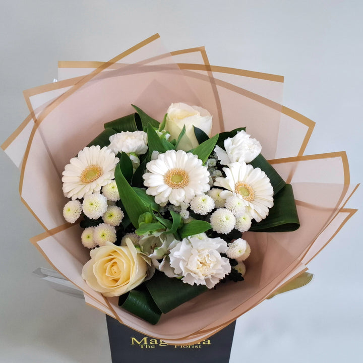 Wadenhoe Bouquet - Magnolia the Florist A pure white hand‑tied bouquet featuring white germini, white bloom carnations, white bloom roses, white alstroemeria, white spray chrysanthemums and gyp. The bouquet is finished in an aqua bubble and then presented in one of our Magnolia‑branded hand‑tied carriers. For scale, our petite size measures approximately 50cm tall and 38cm wide. Larger sizes increase in overall width while maintaining the same balanced proportions.  3