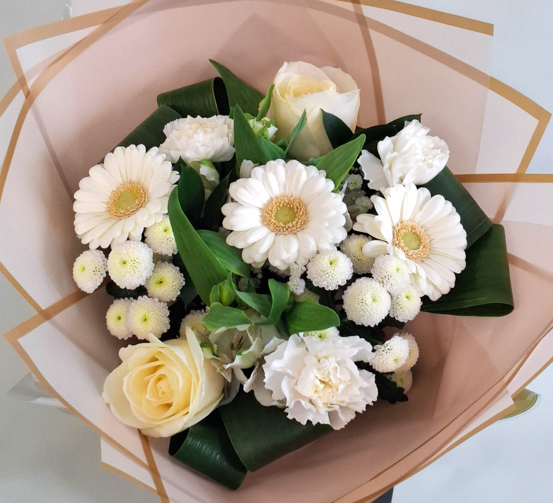 Wadenhoe Bouquet - Magnolia the Florist A pure white hand‑tied bouquet featuring white germini, white bloom carnations, white bloom roses, white alstroemeria, white spray chrysanthemums and gyp. The bouquet is finished in an aqua bubble and then presented in one of our Magnolia‑branded hand‑tied carriers. For scale, our petite size measures approximately 50cm tall and 38cm wide. Larger sizes increase in overall width while maintaining the same balanced proportions.  2