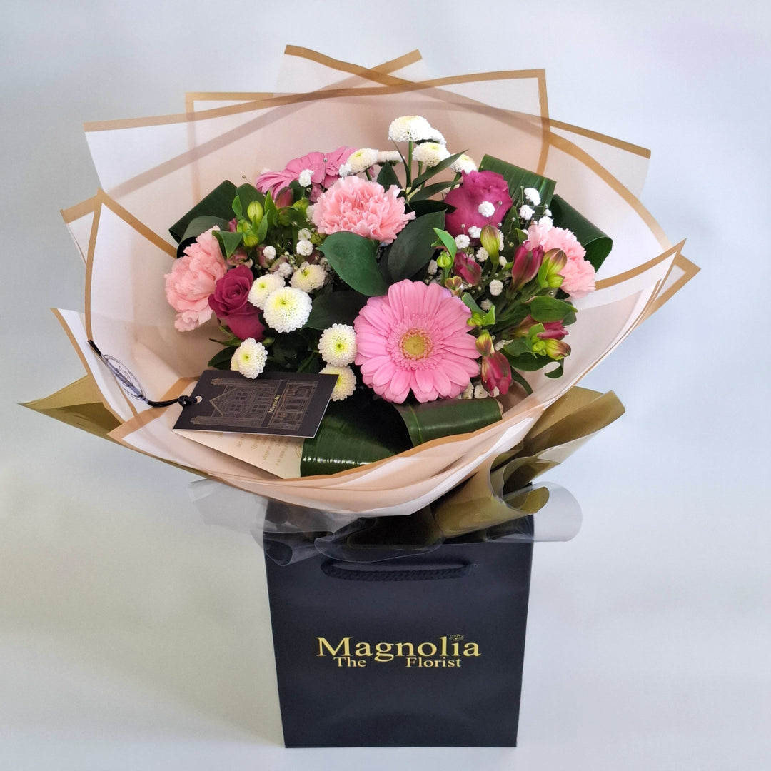 Warkton Bouquet - Magnolia the Florist A bright, feminine hand‑tied bouquet featuring pink alstroemeria, pink carnations, pink germini, pink bloom roses, white spray chrysanthemums, gyp, hard ruscus and folded aspidistra. The bouquet is finished in an aqua bubble and then presented in one of our Magnolia‑branded hand‑tied carriers. For scale, our petite size measures approximately 50cm tall and 38cm wide. Larger sizes increase in overall width while maintaining the same balanced proportions.  1