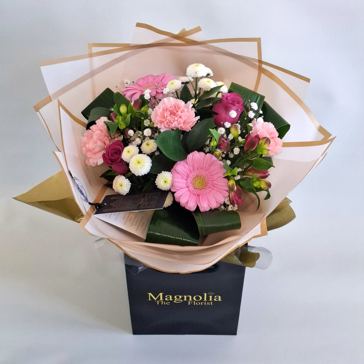 Warkton Bouquet - Magnolia the Florist A bright, feminine hand‑tied bouquet featuring pink alstroemeria, pink carnations, pink germini, pink bloom roses, white spray chrysanthemums, gyp, hard ruscus and folded aspidistra. The bouquet is finished in an aqua bubble and then presented in one of our Magnolia‑branded hand‑tied carriers. For scale, our petite size measures approximately 50cm tall and 38cm wide. Larger sizes increase in overall width while maintaining the same balanced proportions.  3