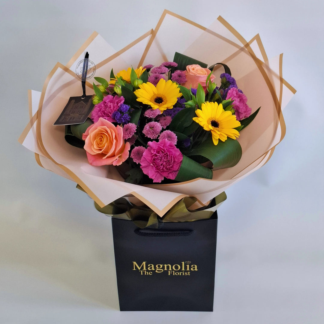 Yarwell Bouquet - Magnolia the Florist A lively, colourful hand‑tied bouquet featuring yellow germini, cerise bloom carnations, orange roses, pink alstroemeria, pink spray chrysanthemums and purple statice. The bouquet is finished in an aqua bubble and then presented in one of our Magnolia‑branded hand‑tied carriers. For scale, our petite size measures approximately 50cm tall and 38cm wide. Larger sizes increase in overall width while maintaining the same balanced proportions.  1