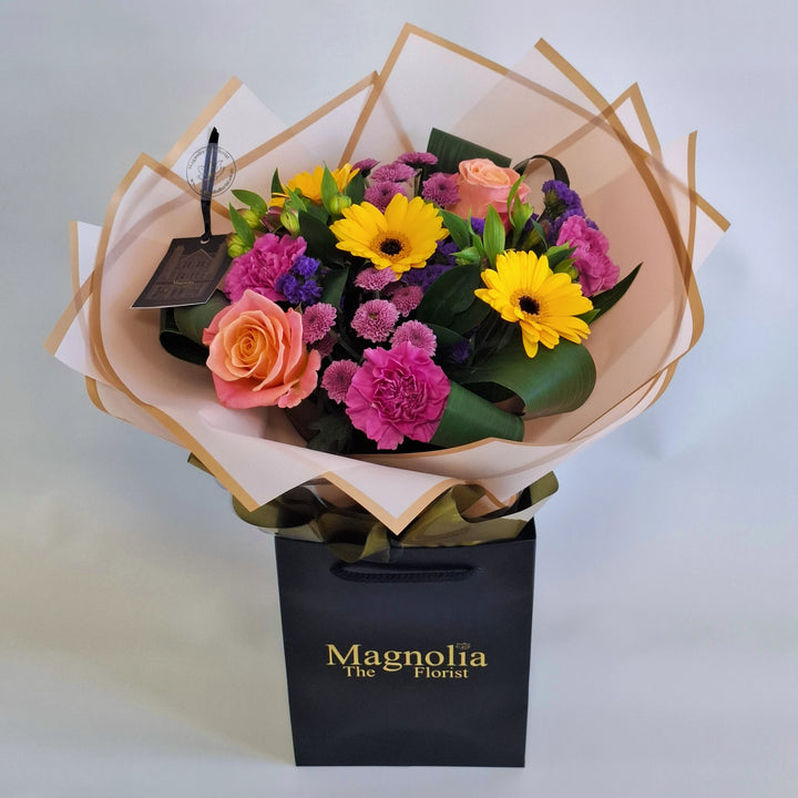 Yarwell Bouquet - Magnolia the Florist A lively, colourful hand‑tied bouquet featuring yellow germini, cerise bloom carnations, orange roses, pink alstroemeria, pink spray chrysanthemums and purple statice. The bouquet is finished in an aqua bubble and then presented in one of our Magnolia‑branded hand‑tied carriers. For scale, our petite size measures approximately 50cm tall and 38cm wide. Larger sizes increase in overall width while maintaining the same balanced proportions.  1