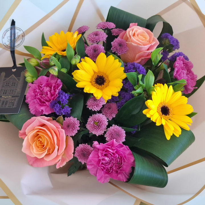 Yarwell Bouquet - Magnolia the Florist A lively, colourful hand‑tied bouquet featuring yellow germini, cerise bloom carnations, orange roses, pink alstroemeria, pink spray chrysanthemums and purple statice. The bouquet is finished in an aqua bubble and then presented in one of our Magnolia‑branded hand‑tied carriers. For scale, our petite size measures approximately 50cm tall and 38cm wide. Larger sizes increase in overall width while maintaining the same balanced proportions.  2