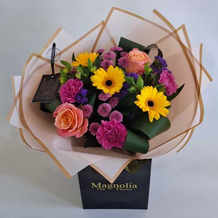 Yarwell Bouquet - Magnolia the Florist A lively, colourful hand‑tied bouquet featuring yellow germini, cerise bloom carnations, orange roses, pink alstroemeria, pink spray chrysanthemums and purple statice. The bouquet is finished in an aqua bubble and then presented in one of our Magnolia‑branded hand‑tied carriers. For scale, our petite size measures approximately 50cm tall and 38cm wide. Larger sizes increase in overall width while maintaining the same balanced proportions.  3